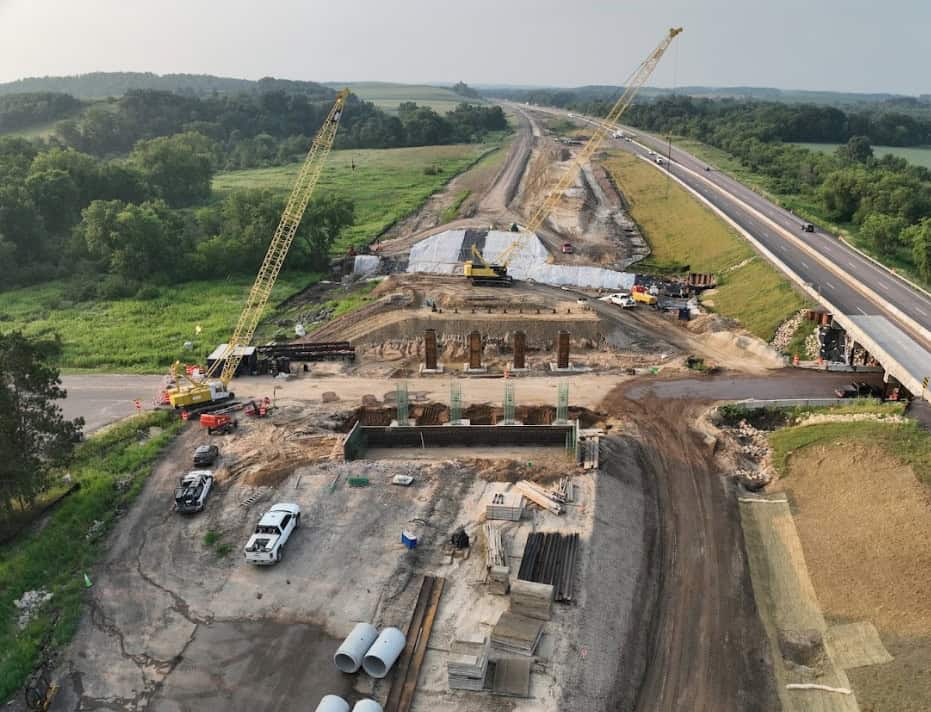 A birds-eye view of construction equipment creating a highway being paved beneath a bridge that is also under construction.